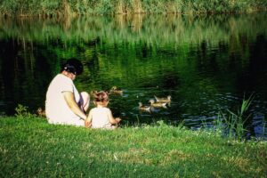 woman sitting with little girl by pond with ducks
